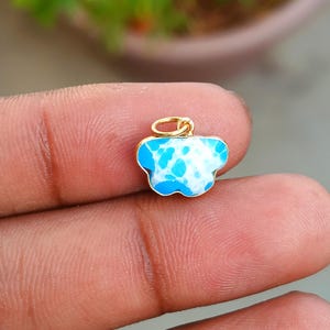 Könnte beinhalten: Ein kleiner, wolkenförmiger Anhänger mit einem goldfarbenen Bügel. Der Anhänger hat ein blau-weiß gesprenkeltes Design mit einem goldfarbenen Rand. Der Schmuck wird in der Hand einer Person gehalten.