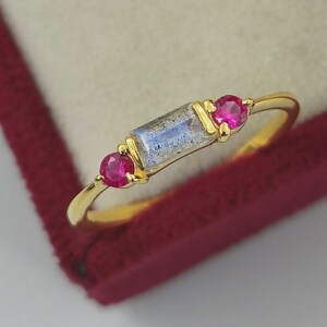 Op de afbeelding: Een gouden ring met een rechthoekige, iriserende edelsteen en twee ronde, roze edelstenen. De ring wordt gepresenteerd tegen een rode fluwelen achtergrond, die het ontwerp en de kleur van het sieraad benadrukt.