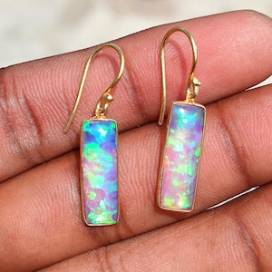 May include: A pair of gold-toned drop earrings featuring rectangular, elongated settings. Each earring contains a vibrant, iridescent opal inlay displaying flashes of bright green, blue, and pink colors. The earrings are attached to simple gold-toned hooks.