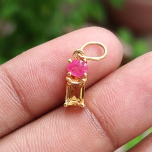 May include: A gold pendant featuring a round, pink gemstone above a rectangular, yellow gemstone. The pendant has a loop at the top for a chain. The gemstones are set in a gold setting.