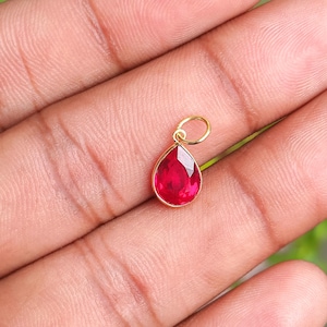 Peut inclure: Pendentif en forme de goutte d'eau avec une pierre précieuse rouge vif sertie dans un cadre en métal doré. La pierre précieuse est facettée, reflétant la lumière. Le pendentif est conçu avec une petite boucle en haut pour l'attacher à une chaîne.