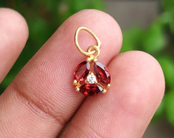 Puede incluir: Un colgante de oro con tres piedras preciosas rojas oscuras ovaladas dispuestas en un patrón triangular alrededor de una pequeña piedra transparente. El colgante tiene un pequeño lazo para una cadena.