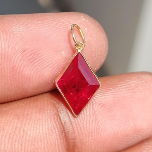 May include: A kite-shaped pendant featuring a faceted, vibrant red gemstone set within a thin gold-toned metal frame. The pendant includes a small, circular gold-toned jump ring at the top for attachment to a chain or cord.