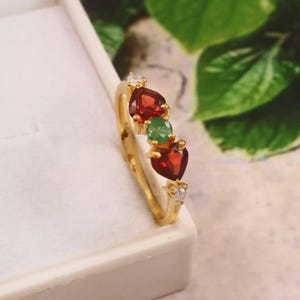 Può includere: Un anello d'oro con pietre rosse a forma di cuore e una pietra verde, rifinito con piccole pietre bianche. L'anello è posto su uno sfondo bianco con fogliame verde sullo sfondo.