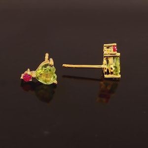 May include: A pair of gold stud earrings with peridot and ruby gemstones. The earrings feature a light green peridot stone and a small red ruby accent. The earrings are set against a dark background.