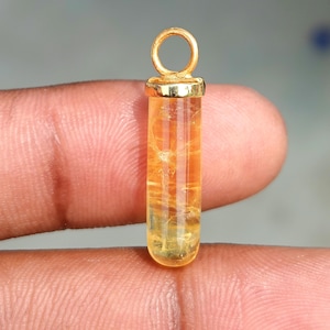 Op de afbeelding: Een cilindervormige hanger met een doorschijnende, goudgele edelsteen met natuurlijke interne insluitingen. De steen is afgewerkt met een gepolijste, goudkleurige metalen kap met een cirkelvormig oog aan de bovenkant voor een ketting.