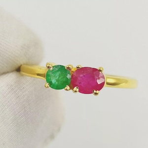Peut inclure: Une bague en or ornée de deux pierres précieuses : une émeraude verte ronde et un rubis rouge foncé ovale. Les pierres sont côte à côte, créant un contraste saisissant. L'anneau est en or poli.