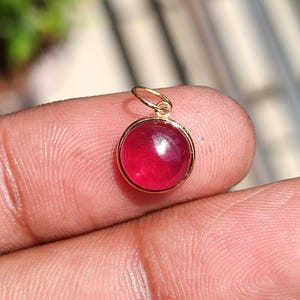 May include: A round, red gemstone pendant with a gold-coloured bezel and loop. The polished gemstone is a vibrant red colour. The pendant is designed to be worn as a necklace charm.