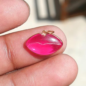 May include: A gold-colored lip-shaped pendant with a bright pink center. The pendant has a small loop at the top for a chain. The lips are outlined in gold and have a subtle white line across the middle.