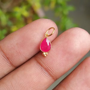 Pode incluir: Um pingente de ouro com uma pedra preciosa vermelha rubi facetada em forma de gota. O pingente tem uma pequena argola na parte superior e uma base dourada decorativa. A pedra preciosa tem cerca de 1,3 cm de comprimento.