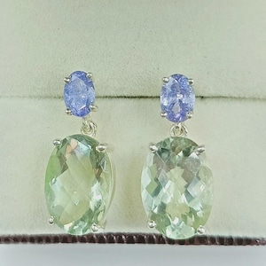 Può includere: Un paio di orecchini pendenti con una gemma ovale verde chiaro sospesa sotto una gemma ovale blu più piccola. Gli orecchini hanno una montatura in metallo color argento e sono esposti su uno sfondo neutro.