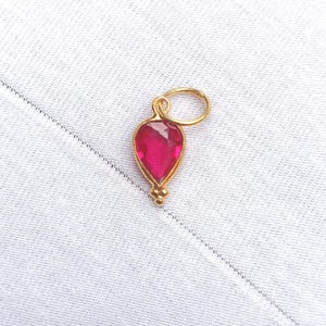 Könnte beinhalten: Ein goldener Anhänger mit einem tropfenförmigen, facettierten, rubinroten Edelstein. Der Anhänger hat oben eine kleine Schlaufe und unten drei kleine goldene Perlen. Der Anhänger ist vor einem weißen Stoffhintergrund platziert.