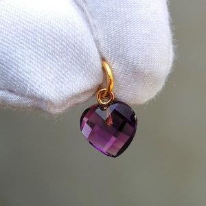 May include: A faceted, heart-shaped amethyst pendant with a gold-colored bail. The deep purple gemstone is cut to reflect light, creating a sparkling effect. The pendant is designed for jewelry making or as a charm.