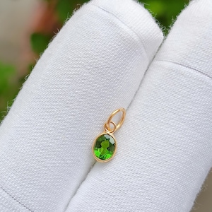 May include: A small, gold pendant featuring a bright green, oval gemstone. The pendant is set in a gold bezel and has a small loop for a chain. The pendant is shown against a white background.