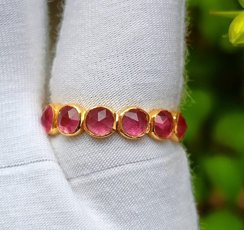 K&ouml;nnte beinhalten: Ein goldener Ring mit einer Reihe von oval geschliffenen, tiefrosa Edelsteinen. Jeder Edelstein ist in einer goldenen Fassung gefasst, wodurch ein durchgehendes Band entsteht. Der Ring wird vor einem wei&szlig;en Stoffhintergrund gezeigt.