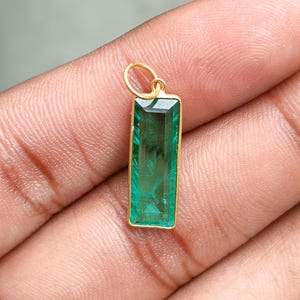 May include: A rectangular pendant featuring a vivid green gemstone within a gold-coloured frame. The gemstone is faceted, and the pendant has a small loop at the top for a chain. The pendant is held in a hand.