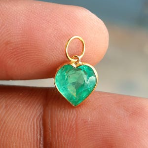 May include: A heart-shaped green gemstone pendant set in a gold frame with a small circular gold jump ring attached at the top. The gemstone features a faceted surface and a vibrant green hue, held by a thin gold metal border.