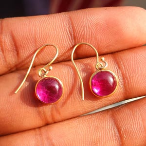 Könnte beinhalten: Ein Paar goldfarbene Ohrringe mit einem runden, magenta-farbenen Edelstein. Die Ohrringe haben ein einfaches Haken-Design und eine kleine, dekorative Kugel über dem Edelstein. Die Edelsteine sind in einer goldenen Lünette gefasst.