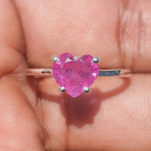 Può includere: Un anello color argento con una pietra preziosa rosa sfaccettata a forma di cuore come pietra centrale. La gemma è fissata da quattro piccole griffe metalliche e la fascia è un design semplice, sottile e in metallo lucido.