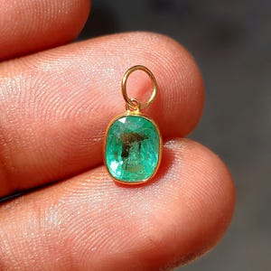 Puede incluir: Un colgante de oro con una piedra preciosa verde de talla esmeralda rectangular. El colgante tiene un pequeño lazo de oro para una cadena. La piedra preciosa está engastada en un bisel de oro.