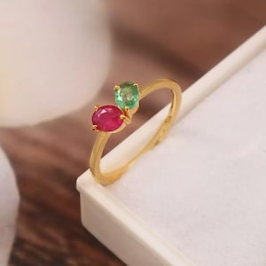 Könnte beinhalten: Ein goldener Ring mit einem Rubin und einem Smaragd. Der Rubin ist oval und leuchtend rot, während der Smaragd ein kleineres Oval in hellem Grün ist. Das Band des Rings ist aus poliertem Gold.