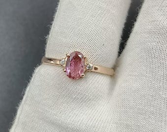 May include: A rose gold ring featuring an oval pink gemstone as the centerpiece, flanked by two smaller, round, clear gemstones. The ring is set on a white surface.