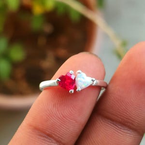 May include: A silver ring featuring two heart-shaped gemstones. One is a vibrant red, the other a shimmering white. The ring's band is thin and delicate, with the hearts set close together, creating a romantic and elegant design.