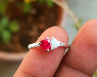 May include: A silver ring featuring two heart-shaped gemstones. One is a vibrant red, the other a shimmering white. The ring's band is thin and delicate, with the hearts set close together, creating a romantic and elegant design.