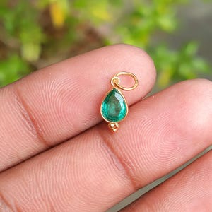 May include: A gold pendant with a pear-shaped, emerald-green gemstone. The pendant has a small loop at the top and a cluster of three small gold beads at the bottom. The gemstone is set in a gold bezel.