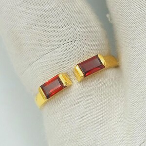 May include: A gold ring featuring two rectangular, deep red gemstones. The ring has an open design, with the gemstones set on either side. The gemstones are approximately 1.3 cm in length.