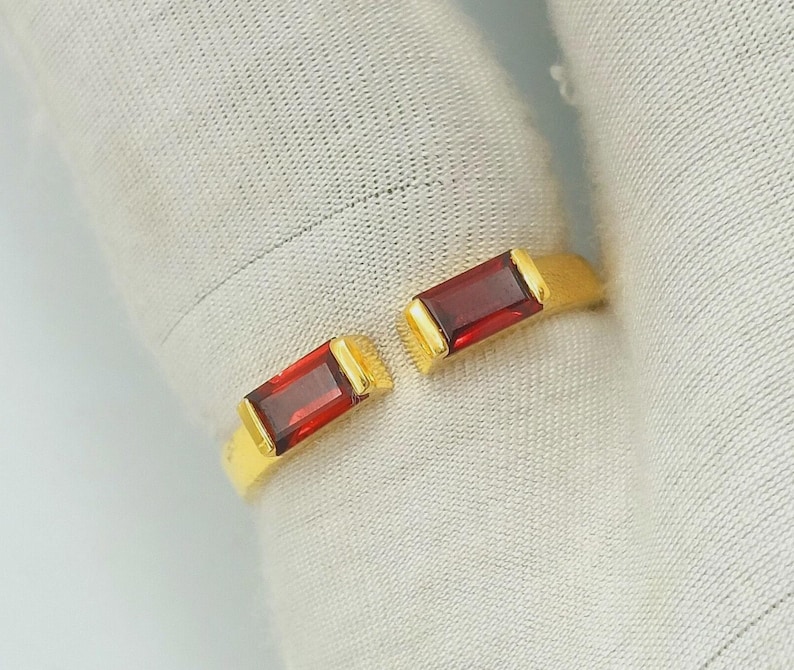 May include: A gold ring featuring two rectangular, deep red gemstones. The ring has an open design, with the gemstones set on either side. The gemstones are approximately 1.3 cm in length.