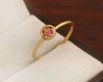 May include: A gold ring with a round, peach-colored gemstone set in a bezel setting. The ring is displayed in a black jewelry box with a white interior. The band is thin and delicate.