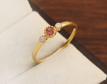 May include: A gold ring with a central round pink gemstone, flanked by two smaller white gemstones. The ring is set in a simple, elegant design, perfect for everyday wear. The ring is displayed in a jewelry box.