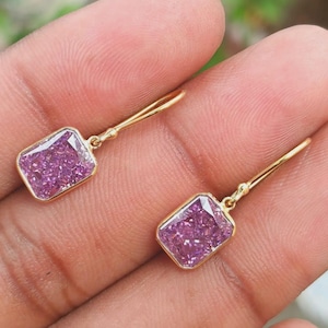 May include: A pair of gold drop earrings featuring rectangular-cut, vibrant purple-pink stones set in polished gold bezels. The earrings are suspended from simple gold hooks, showcasing a faceted surface that reflects light across the stones.
