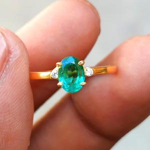 May include: A gold ring featuring an oval-cut, vibrant green gemstone as the centerpiece. Two smaller, clear gemstones flank the main stone. The ring is held between fingers, showcasing its polished gold band and the brilliance of the gemstones.