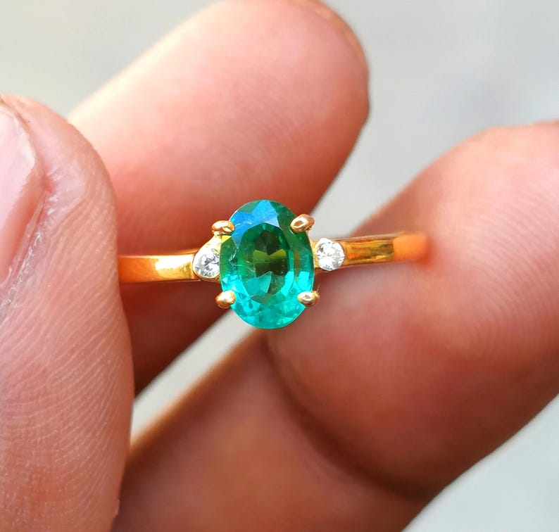 May include: A gold ring featuring an oval-cut, vibrant green gemstone as the centerpiece. Two smaller, clear gemstones flank the main stone. The ring is held between fingers, showcasing its polished gold band and the brilliance of the gemstones.