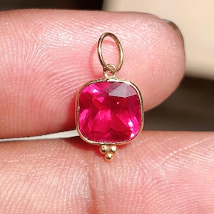 Pode incluir: Um pingente de ouro com uma pedra preciosa de corte quadrado em um tom vibrante de vermelho rosado, engastada em uma moldura de ouro polido. O pingente inclui um pequeno aro de ouro no topo e um detalhe decorativo de três pequenas esferas.