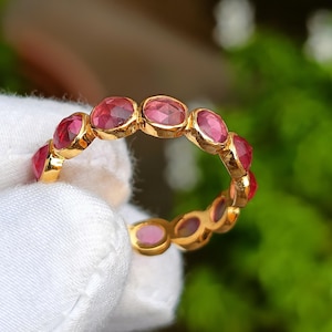 Könnte beinhalten: Ein goldener Ring mit einer Reihe von oval geschliffenen, tiefrosa Edelsteinen. Die Edelsteine sind eng aneinander gesetzt und bilden ein durchgehendes Farbband. Das Goldband des Rings bildet einen warmen Kontrast zu den leuchtend rosa Steinen.