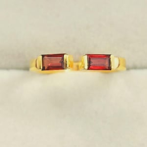May include: A gold ring with two rectangular, deep red gemstones. The ring has a simple, elegant design with the gemstones set side-by-side. The gold band provides a warm contrast to the rich colour of the stones, creating a striking piece of jewellery.