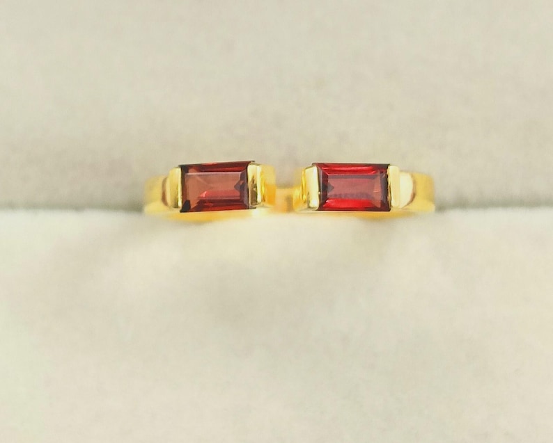 May include: A gold ring with two rectangular, deep red gemstones. The ring has a simple, elegant design with the gemstones set side-by-side. The gold band provides a warm contrast to the rich colour of the stones, creating a striking piece of jewellery.