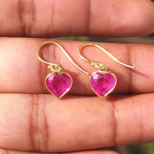 May include: A pair of gold heart-shaped earrings, each featuring a bright pink gemstone. The earrings are presented against a skin-toned background, highlighting their design and colour.