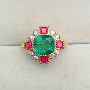 Puede incluir: Un anillo de oro con una gran esmeralda central de corte cuadrado, rodeada de piedras rojas cuadradas más pequeñas y diamantes blancos. El anillo tiene un diseño clásico y ornamentado.