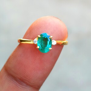 May include: A gold ring featuring an oval-cut emerald gemstone, flanked by two smaller white gemstones. The ring is set on a finger, showcasing the vibrant green of the emerald against a blurred background. A classic piece of jewelry.