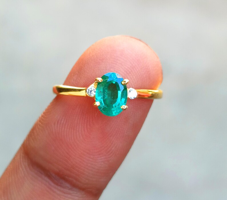 May include: A gold ring featuring an oval-cut emerald gemstone, flanked by two smaller white gemstones. The ring is set on a finger, showcasing the vibrant green of the emerald against a blurred background. A classic piece of jewelry.