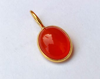 May include: A gold pendant featuring a bright red, oval gemstone. The gemstone is set within a gold bezel and has a polished, reflective surface. The pendant has a small gold loop at the top for a chain.