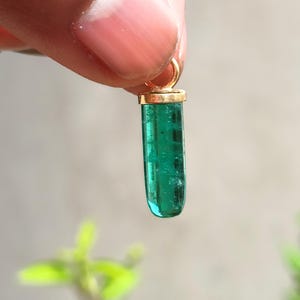 Op de afbeelding: Een felgroene edelsteen hanger met een goudkleurige dop en lus. De smaragdgroene steen is cilindrisch en lijkt een natuurlijke kristal te zijn. De hanger wordt tegen een vage achtergrond gehouden.