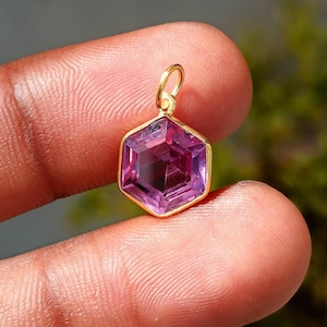 May include: A hexagonal pendant featuring a faceted, deep purple gemstone set within a gold-coloured bezel. The pendant has a small loop at the top for a chain. The gemstone is held between two fingers.