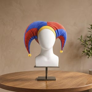 May include: A jester hat featuring a multi-colored design with alternating red and blue fabric panels. The hat includes a bright yellow band around the base and two long, curved points that hang down on each side, each tipped with a small yellow pom-pom.