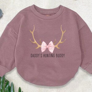 May include: A mauve-coloured sweatshirt with long sleeves and ribbed cuffs. The front displays gold antlers, a pink bow, and the text "DADDY'S HUNTING BUDDY". The image has a festive theme, with Christmas decorations in the background.