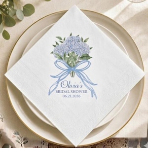 May include: A white square paper napkin featuring a watercolour illustration of a blue hydrangea bouquet tied with a light blue ribbon. The text reads Olivia's BRIDAL SHOWER 06.21.2026 in blue lettering, centred below the floral design.